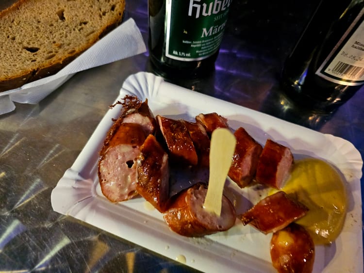 An unrelated image of a Käsekrainer on a paper plate, with a piece of bread and a bottle of beer